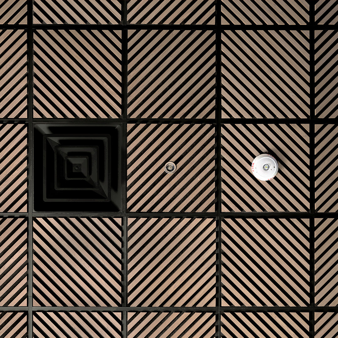 A close-up of a modern wall or ceiling panel featuring a grid pattern of square tiles with diagonal slats in a warm brown tone, separated by black grout lines. One tile has a concentric square design, and another has a small circular metallic fixture.
