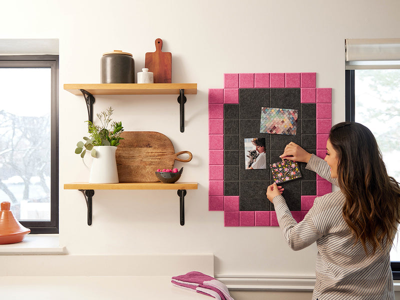 Woman arranging photos on a decorative wall panel with pink and black tiles in a kitchen.