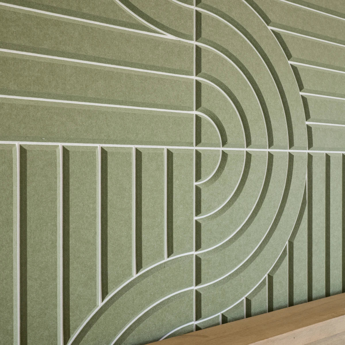 A close-up of a textured olive green wall panel with a raised geometric pattern of concentric arches and vertical lines, with a light wood surface visible at the bottom edge.