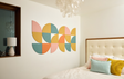 A Felt Right Wall Design quarter circle pieces in shades of pink, yellow, orange, and blue on a white wall in a bedroom next to a window with a lamp and a bed.