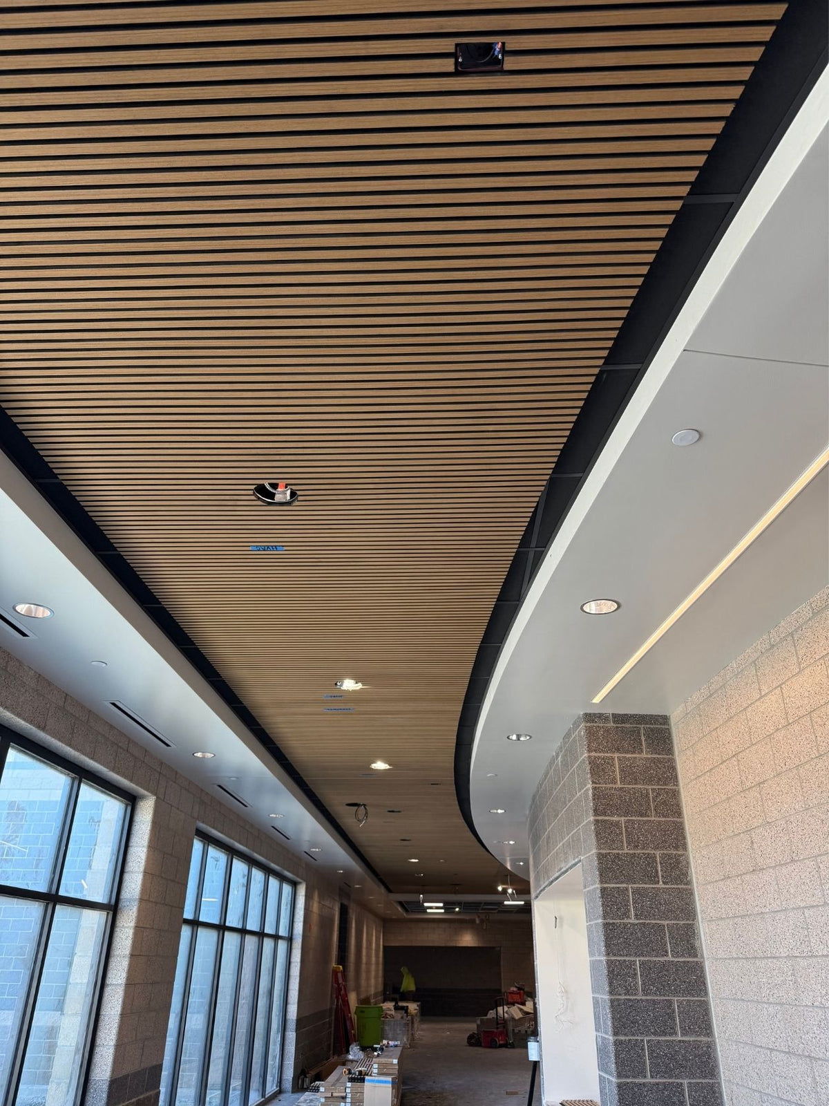 Wood slat ceiling panels installed in a modern indoor corridor with stone columns, recessed lighting, and large windows.