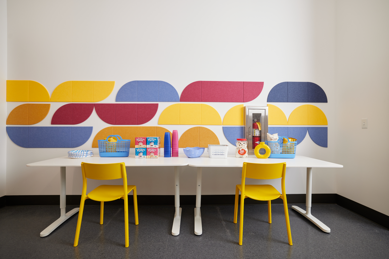 A Felt right wall art design in shades of pink, yellow, and blue tiles on a white wall with a table and two chairs.