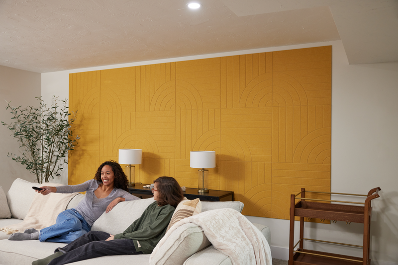 A Felt Right Wall design in a yellow shade in a living room behind a table with two lamps and a couch with two people on it.