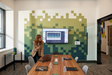 A Felt Right wall design in a conference room along the wall in a pixelated design in shades of green around a TV and a lady leading a discussion.