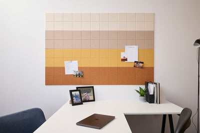A Felt Right wall design bulletin board with a gradient of colors ranging from beige to ochre, mounted on a wall above a white desk with various items such as a photo frame, a potted plant, and a closed laptop.