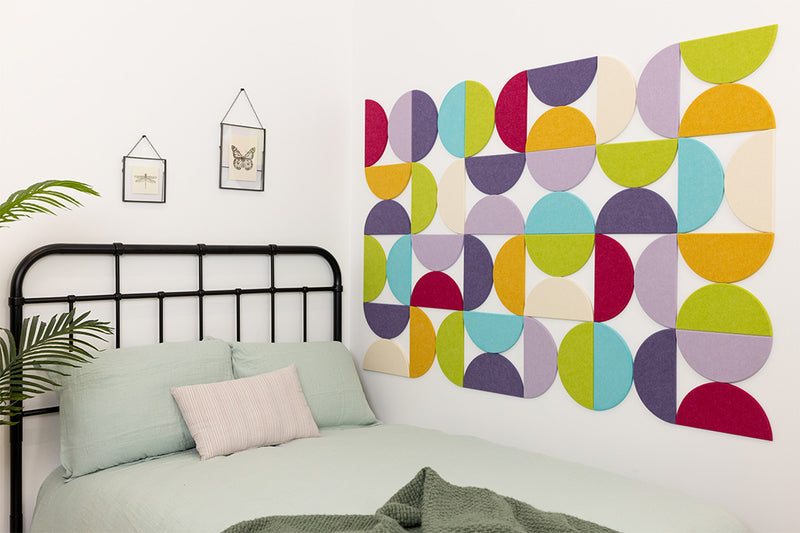 A Felt Right wall design using half circles in the shades of purple, blue, yellow, green, and cream on a wall next to a bed.