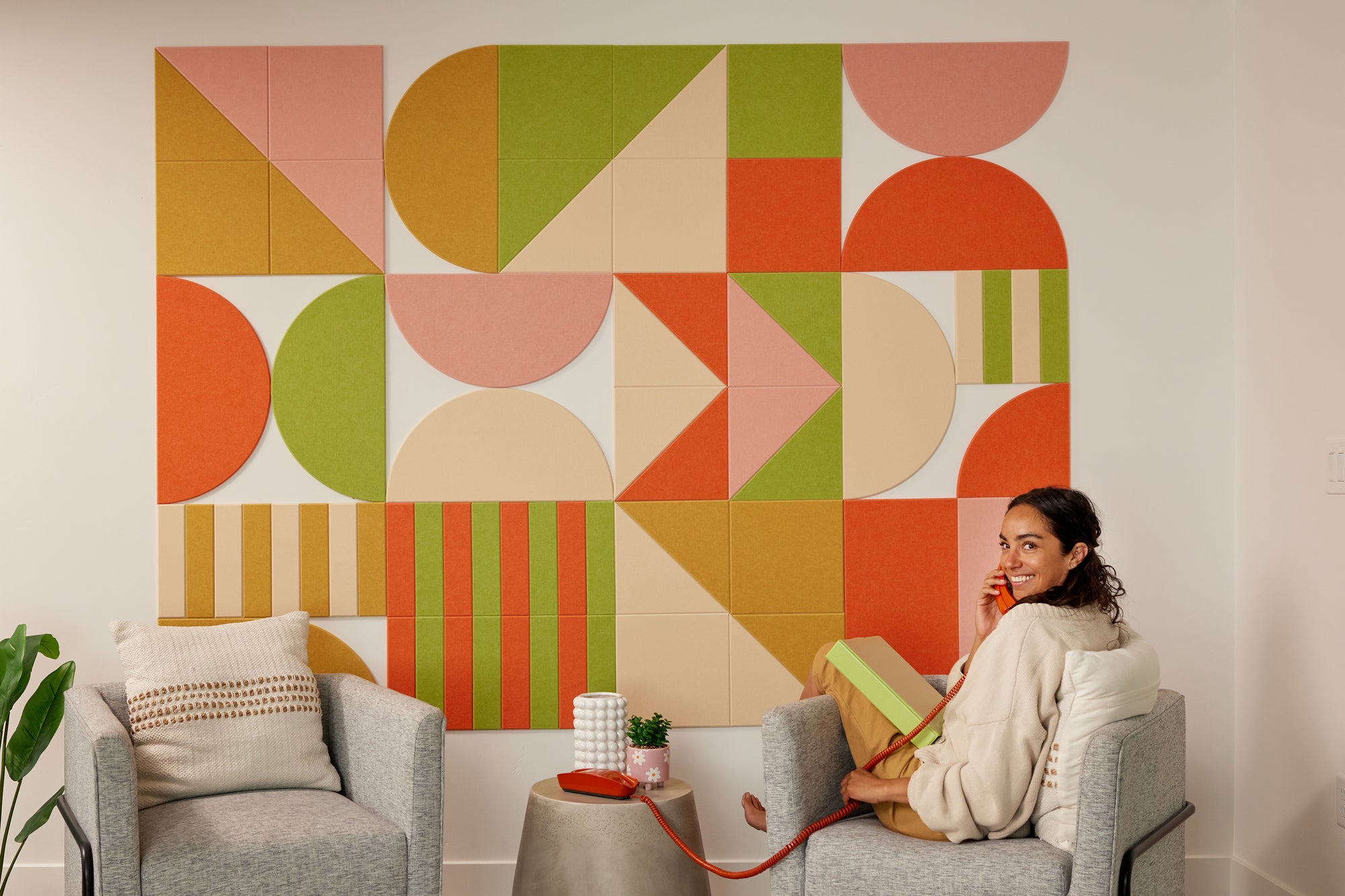 A Felt Right wall art design using tripes and half circles in shades of pink, green, and yellow. in a living room with two chairs and a woman talking on the phone sitting in a chair.