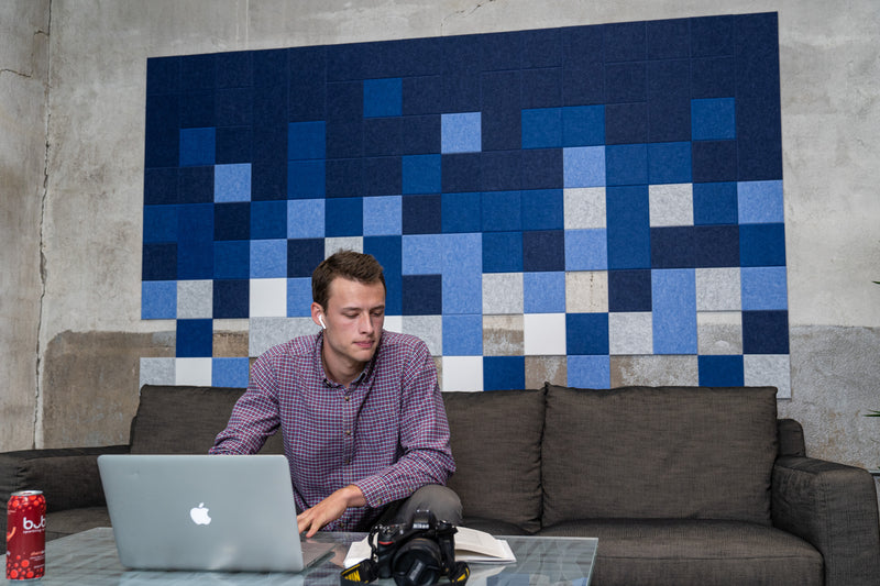 A Felt Right wall art design using shades of blue, white and grey small square felt tiles to create a pattern on a cement wall with a man working on his computer while sitting on a couch.