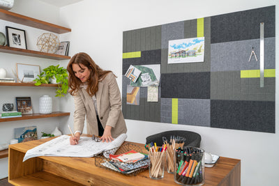 A Pinnable Felt Right wall art design in shades of grey and green felt tiles on a white wall in a n office with a woman working at the desk.