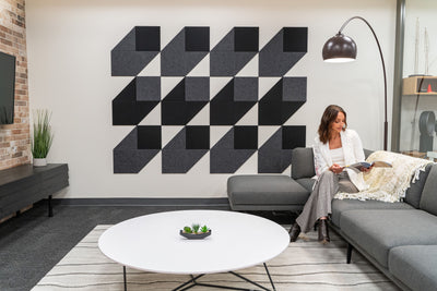 A Felt Right Wall art design in shades of grey giving a 3d vibe of pillars on a wall next to a couch with a women reading on it.