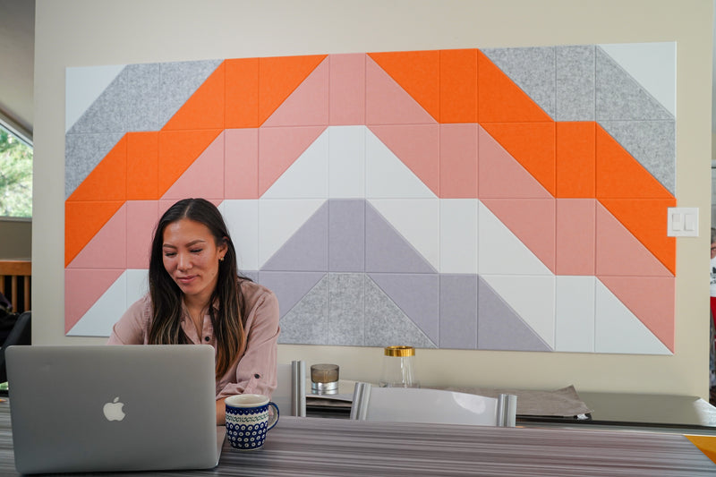 A Felt Right wall art design in shades of grey, orange, pink, and purple making a geometric summit pattern on felt tiles on a wall with a woman working at her desk.