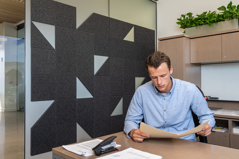 A Felt Right wall art design in shade of black felt tiles on a wall in an office with a man working at his desk.