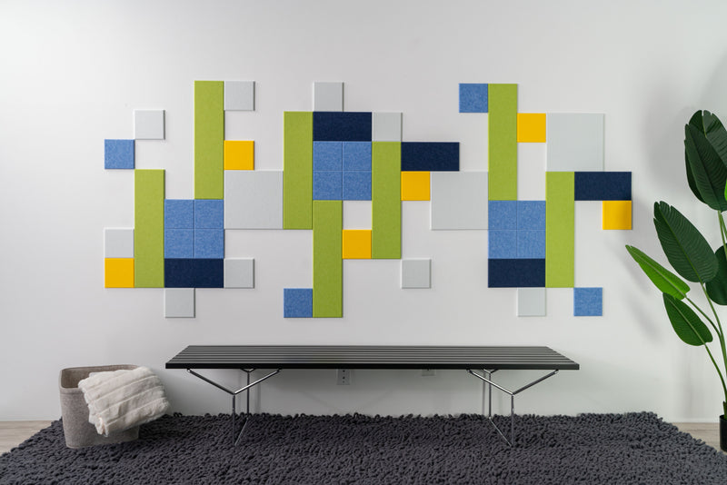 a Felt tight wall art design in shades go green, blue,  yellow and white felt tiles on a white wall above a bench on a rug.