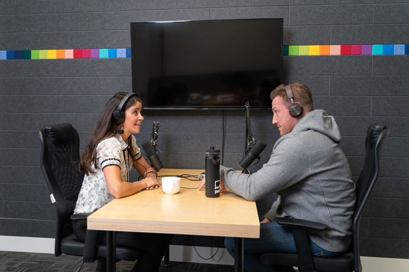 A Felt right wall art design in in a dark grey with a rainbow colored strip in a room wit people doing a podcast.