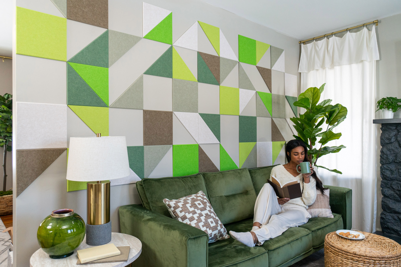 A Felt Right wall art design in shades of green, brown, and white creating a backdrop behind a couch and a side table with a lamp.