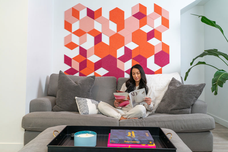 A Felt Right art wall design in shades of pink and orange creating 3d-like images in a pattern on a wall in a living room behind a could with a woman reading a magazine.