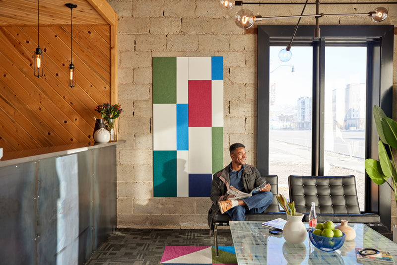 A Felt Right wall art design in shades of blue, green, red, and cream in a geometric design on a brick wall in an office with two chairs and a big window.
