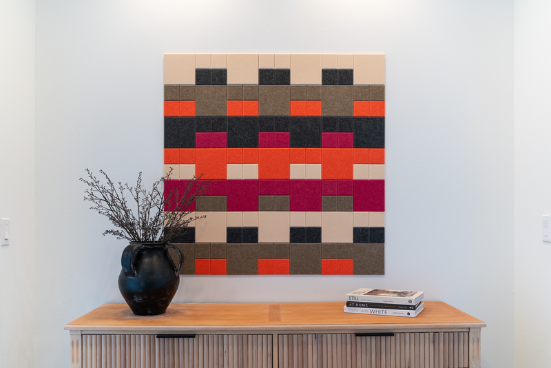 A Felt Right wall design in shades of cream, brown, black, orange, and pink creating a beautiful display above side table that has a plant and books on it.