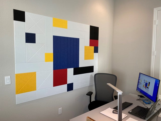 A Felt Right wall art design in white, yellow, blue, and black felt tiles on a wall in an office next to a desk with a computer on it.
