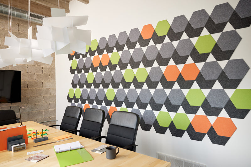 A Felt Right wall art design in shades of grey, orange, and green in a hexagon shapes on a white wall in a conference room or an office.