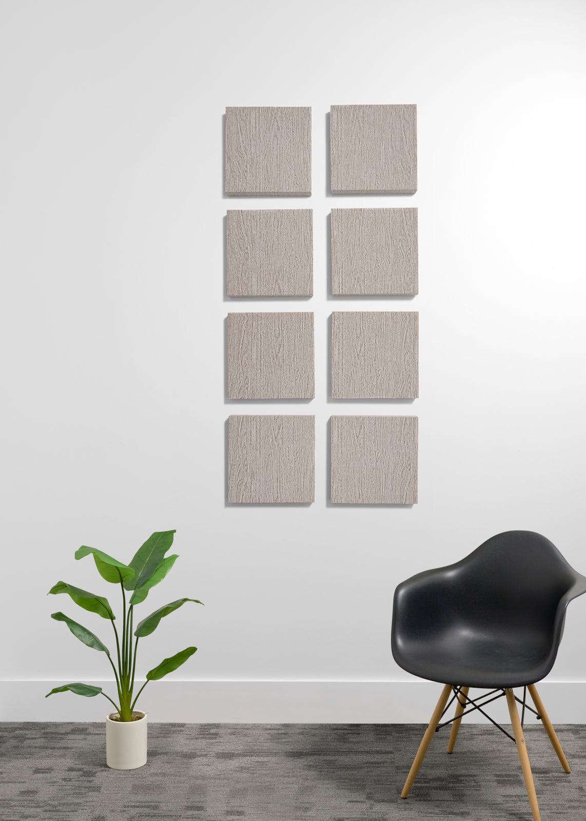 Eight square wooden wall panels with a natural wood grain finish are arranged in a grid on a white wall. The scene is styled with a black molded plastic chair with wooden legs on the right and a green potted plant on the left, set against a gray textured floor.
