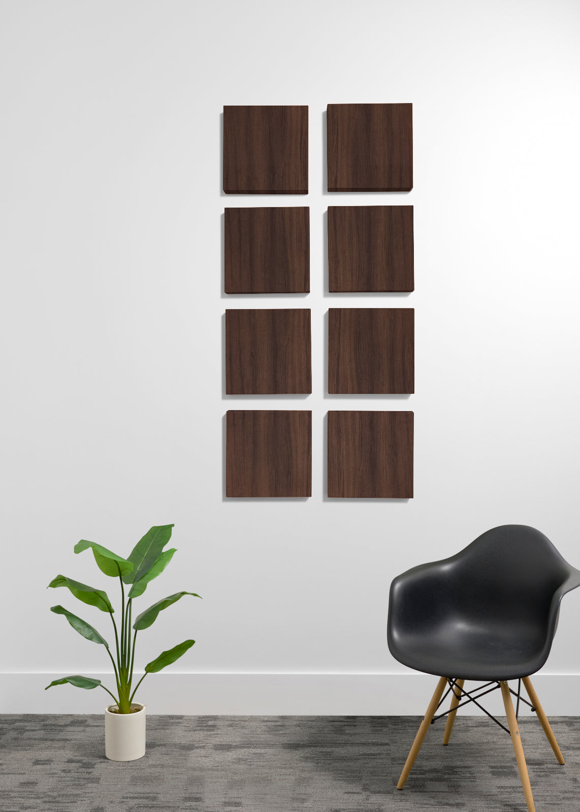 Eight square wooden wall panels with a natural wood grain finish are arranged in a grid on a white wall. The scene is styled with a black molded plastic chair with wooden legs on the right and a green potted plant on the left, set against a gray textured floor.