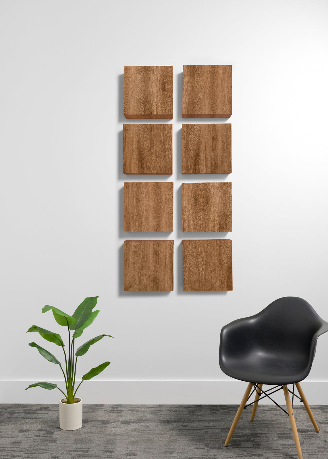Eight square wooden wall panels with a natural wood grain finish are arranged in a grid on a white wall. The scene is styled with a black molded plastic chair with wooden legs on the right and a green potted plant on the left, set against a gray textured floor.