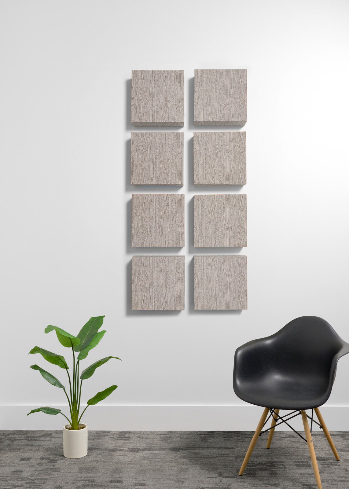 Eight square wooden wall panels with a natural wood grain finish are arranged in a grid on a white wall. The scene is styled with a black molded plastic chair with wooden legs on the right and a green potted plant on the left, set against a gray textured floor.