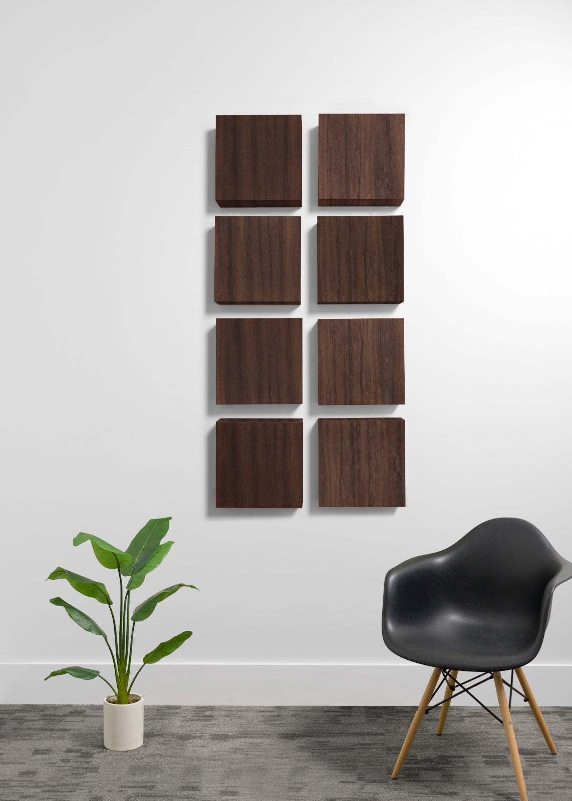 Eight square wooden wall panels with a natural wood grain finish are arranged in a grid on a white wall. The scene is styled with a black molded plastic chair with wooden legs on the right and a green potted plant on the left, set against a gray textured floor.