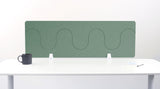 Green desk divider with wavy cutout pattern mounted on white desk with supports.