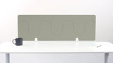 Sage green desk divider with wavy cutout pattern mounted on white desk with metal brackets.