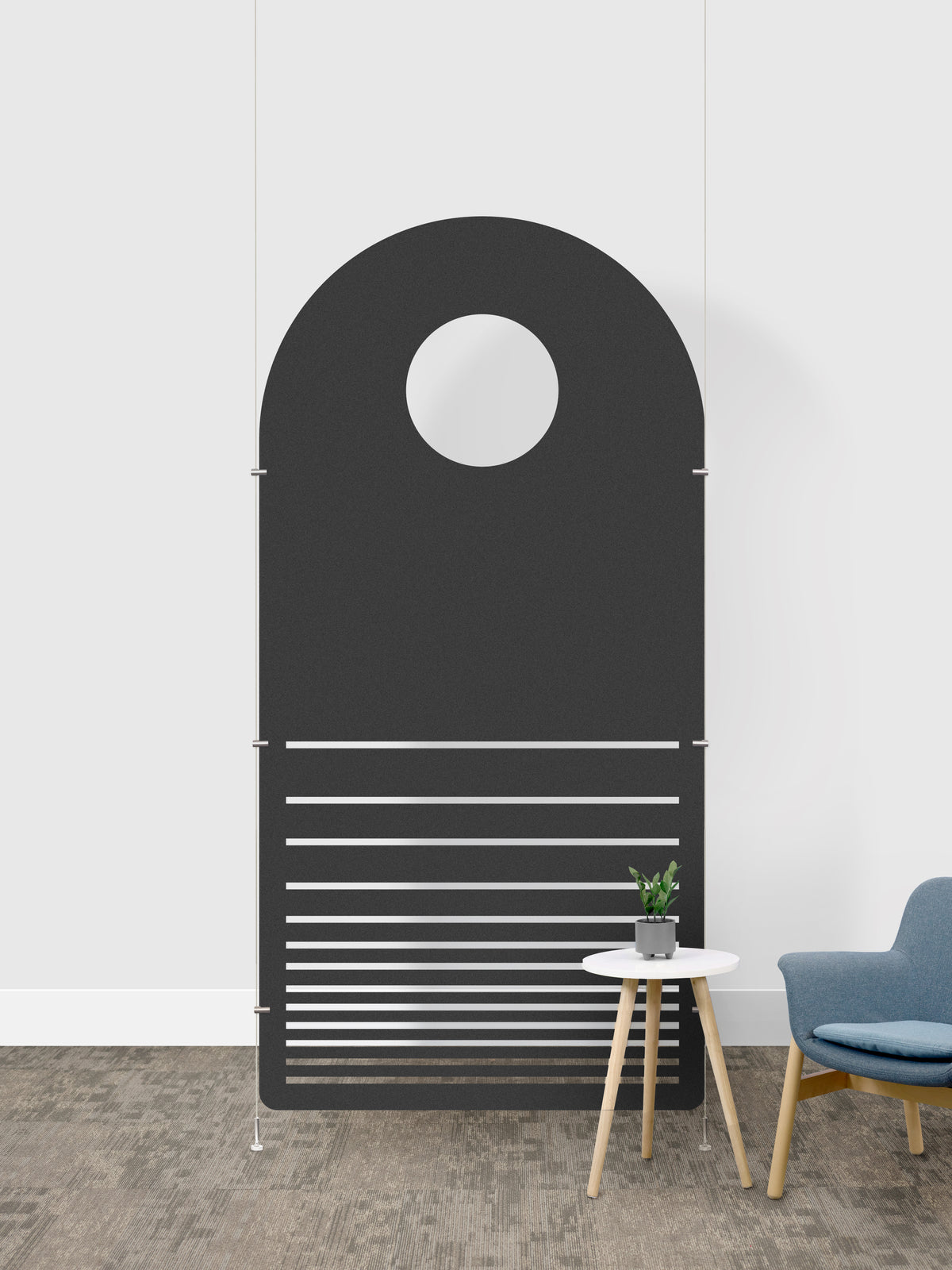A minimalist arched room divider in a dark gray color, featuring a circular cutout at the top and horizontal slats on the lower half. The divider is shown in a modern room setting with a small white side table, a potted plant, and a blue armchair against a white wall.