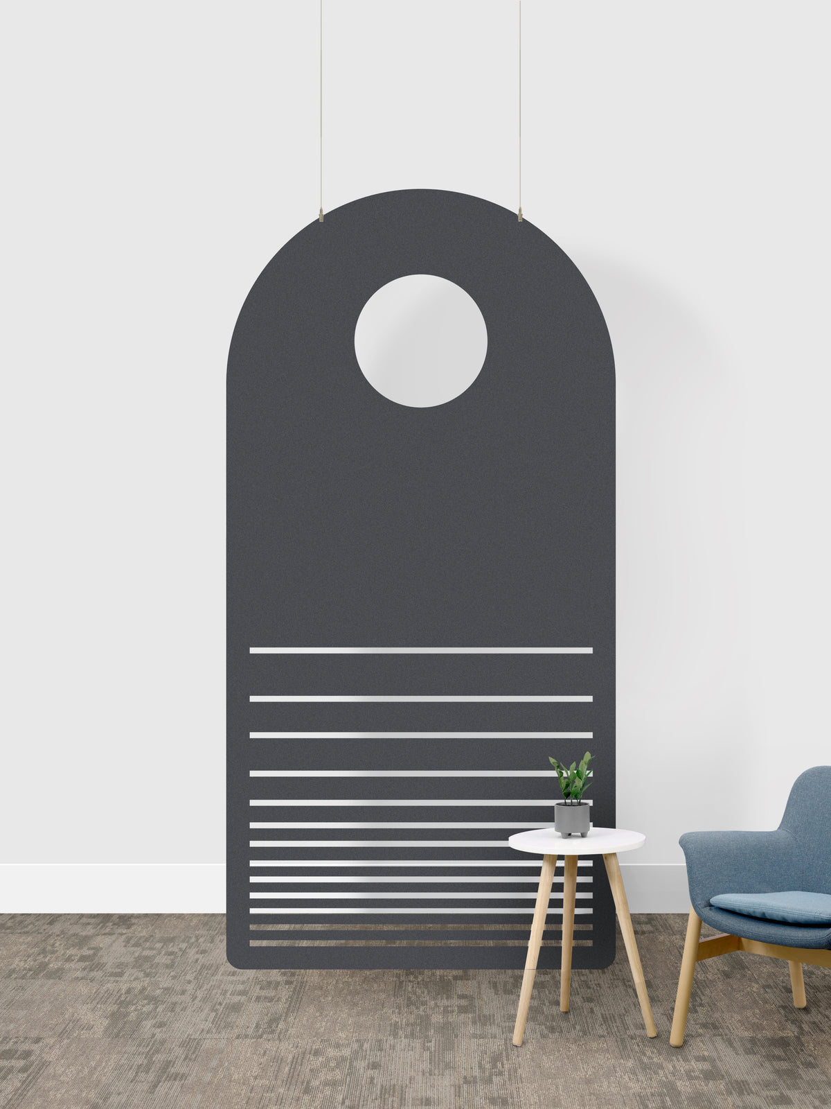 A modern dark gray arched wall art piece with a circular cutout near the top and horizontal slat details near the bottom, shown hanging in a minimalist room setting next to a small side table and blue chair.