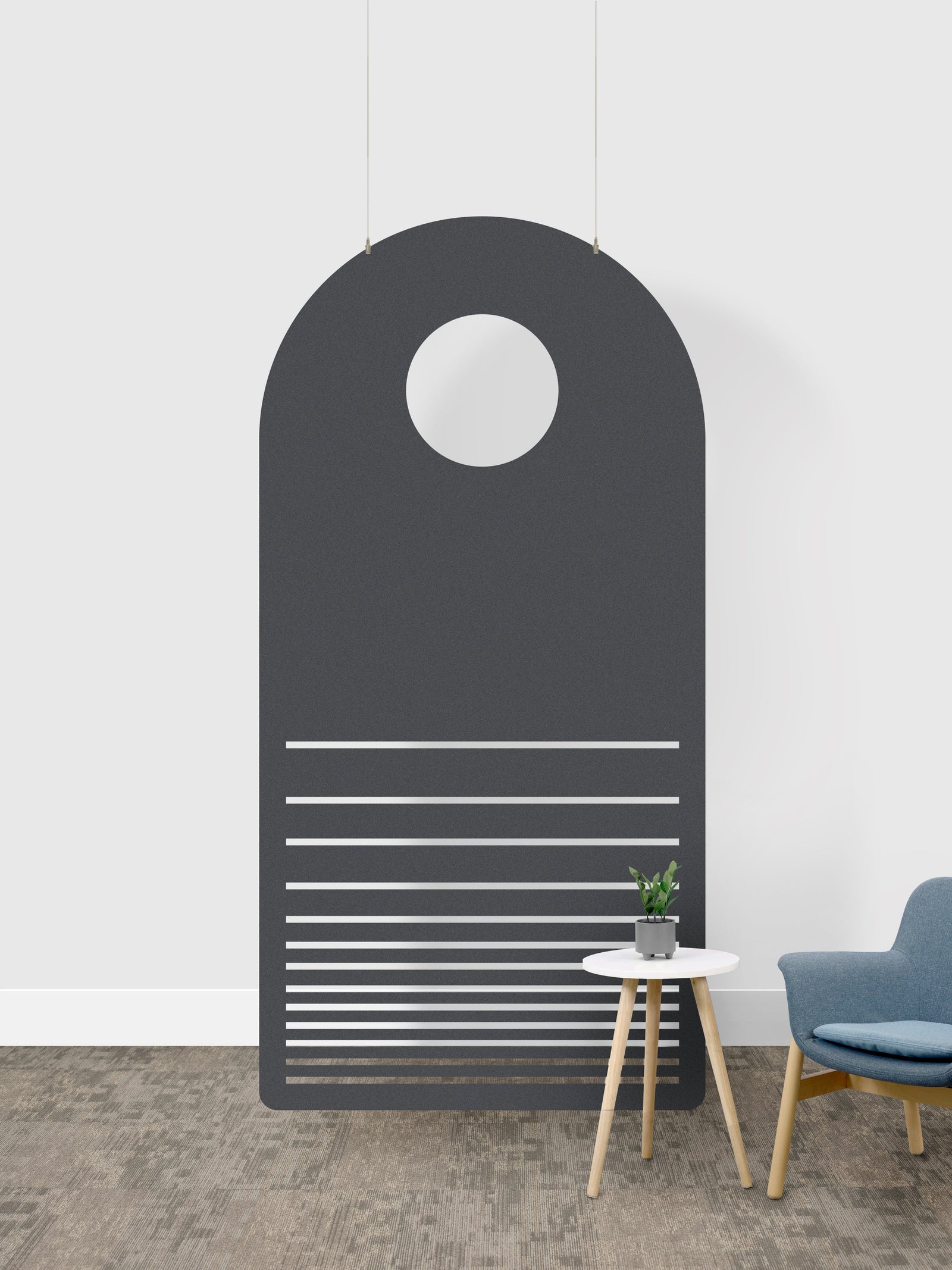 A modern dark gray arched wall art piece with a circular cutout near the top and horizontal slat details near the bottom, shown hanging in a minimalist room setting next to a small side table and blue chair.