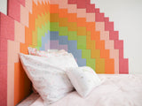 Felt Right wall art design using pixel square tiles in shades of the rainbow to create a pixel look of the rainbow on a corner of two walls in a bedroom next to a bed.