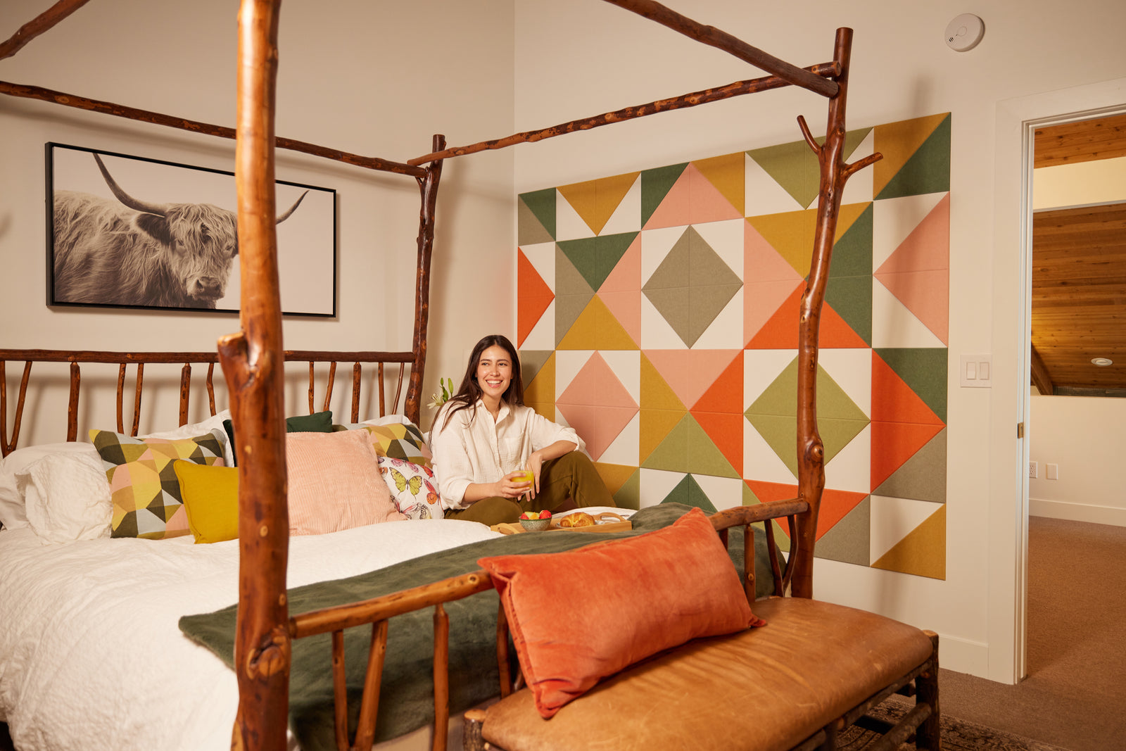 A Felt Right wall art design next to a bed in shades of pink, green, orange, and grey in a bedroom with cow art on the wall and a wooden bed with a women sitting on it.