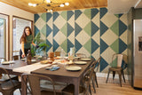 A Felt Right wall art design in shades of green and cream triangles tiles covering a wall in a dinning room with a women setting the dinning table.