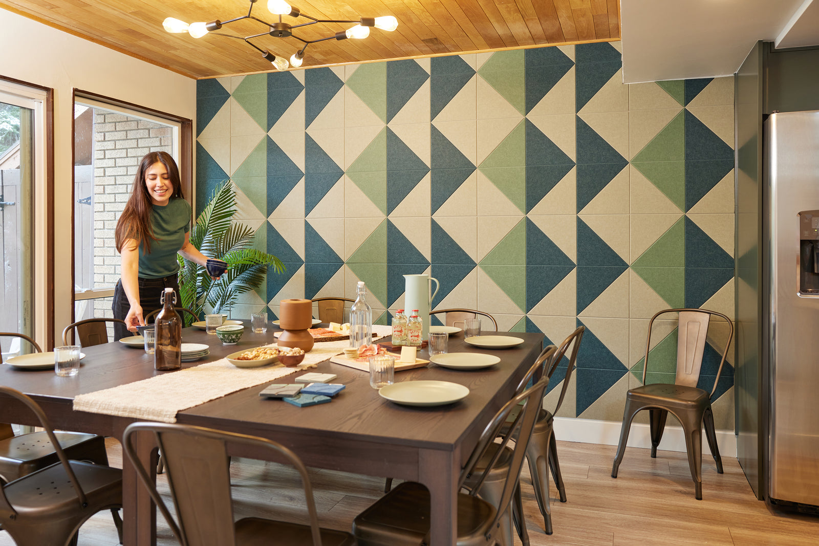 A Felt Right wall art design in shades of green and cream triangles tiles covering a wall in a dinning room with a women setting the dinning table.