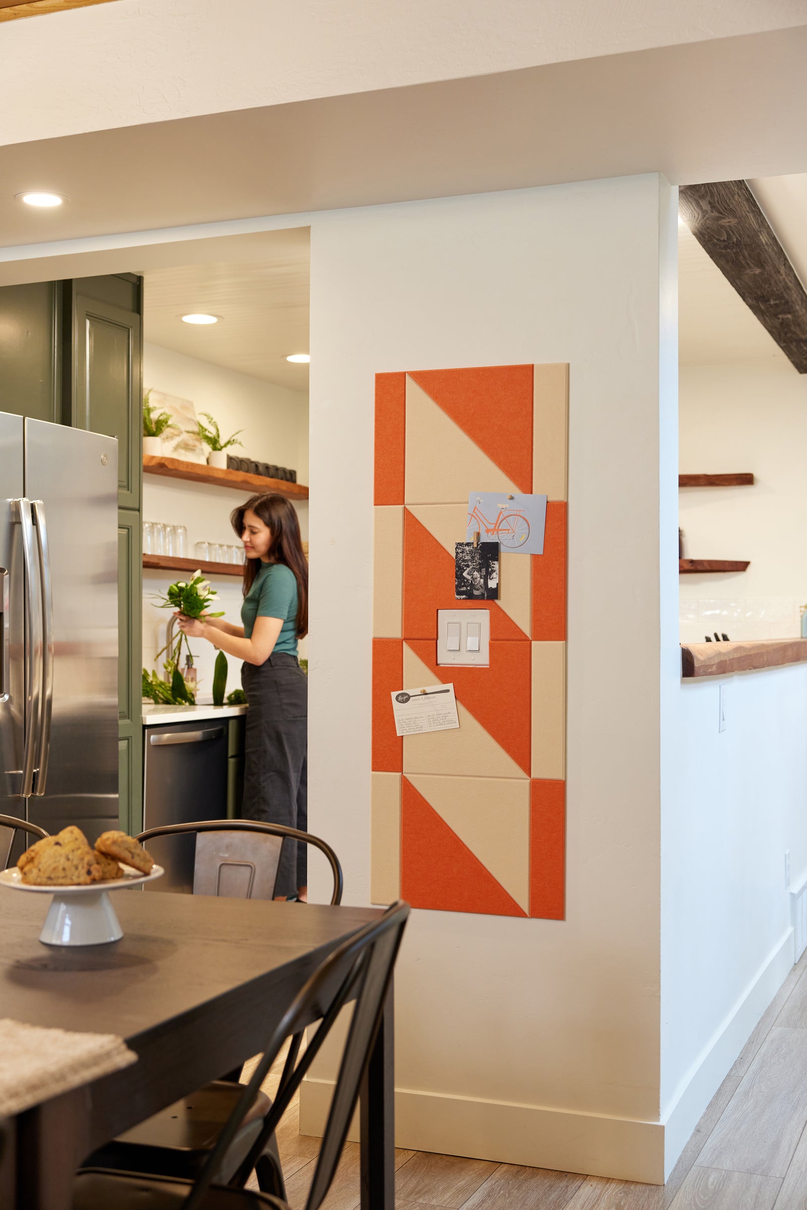 A Felt Right modern design  orange and white geometric designed wall decor or panel system located on a wall in a home environment, with a woman in the background appearing to be cooking in the kitchen.