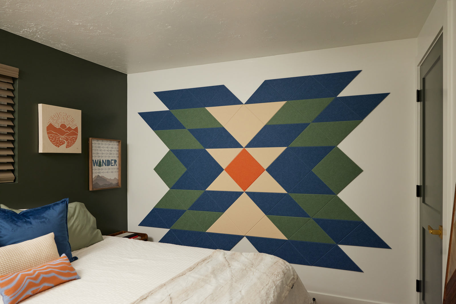 A Felt Right wall art design using shades of blue, green, cream, and orange triangle felt tiles on a white wall in a bedroom next to a bed.