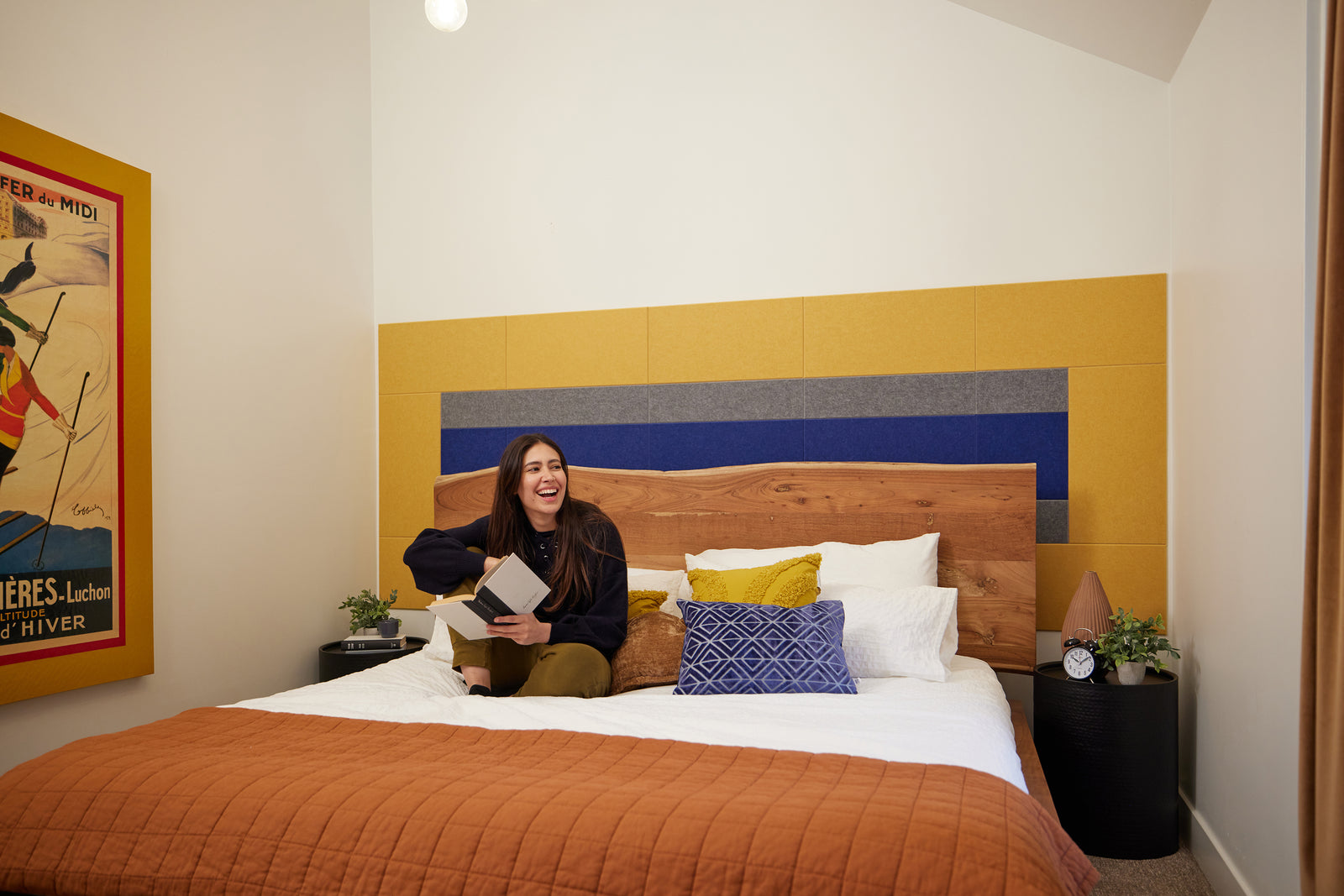 A Felt Right wall design in blue yellow and grey felt tiles on a white wall in a bedroom behind a bed with a girl reading on it 