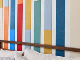 Felt right wall art design in a bedroom using shades of red, orange, blue, and cream felt tiles on a white wall and ceiling behind a bed.