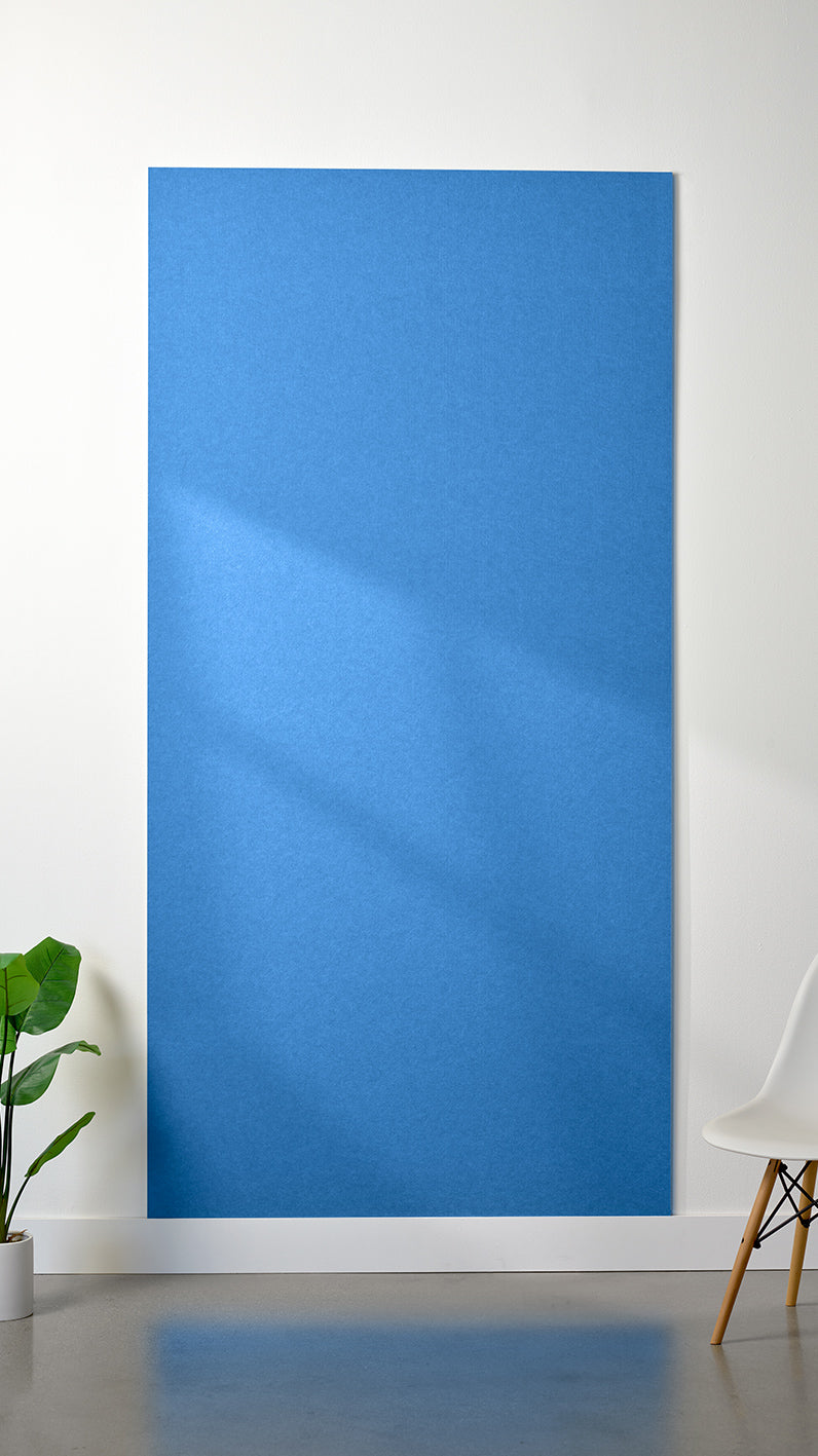 Bright blue Felt Right 4’ x 8’ PET felt panel with no engraving against a white wall. A plant and chair are in the foreground.