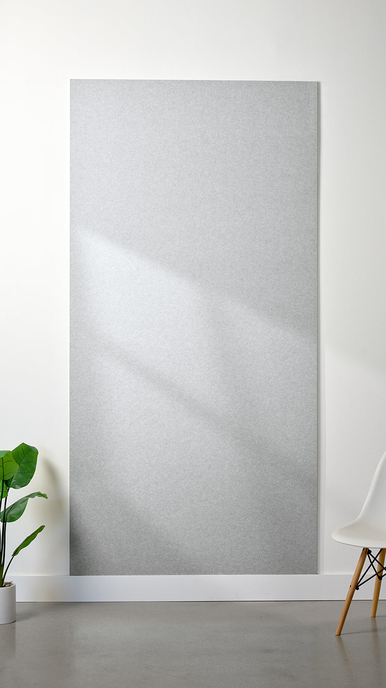 Heathered light grey Felt Right 4’ x 8’ PET felt panel with no engraving against a white wall. A plant and chair are in the foreground.