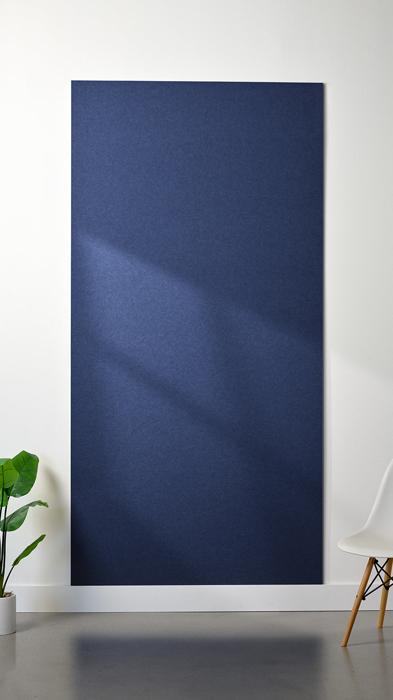 Dark blue Felt Right 4’ x 8’ PET felt panel with no engraving against a white wall. A plant and chair are in the foreground.