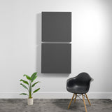Two stacked dark gray square acoustic panels mounted on white wall above chair and plant.