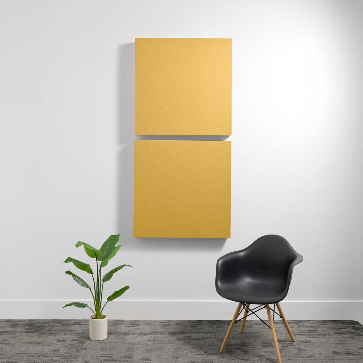 Two stacked mustard-yellow square acoustic panels mounted on white wall above black chair and potted plant.
