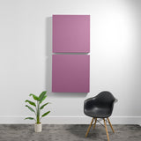 Two mauve square acoustic panels mounted vertically on a white wall above a black chair and potted plant.