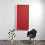 Two red square acoustic panels mounted vertically on a white wall above a black chair and potted plant.