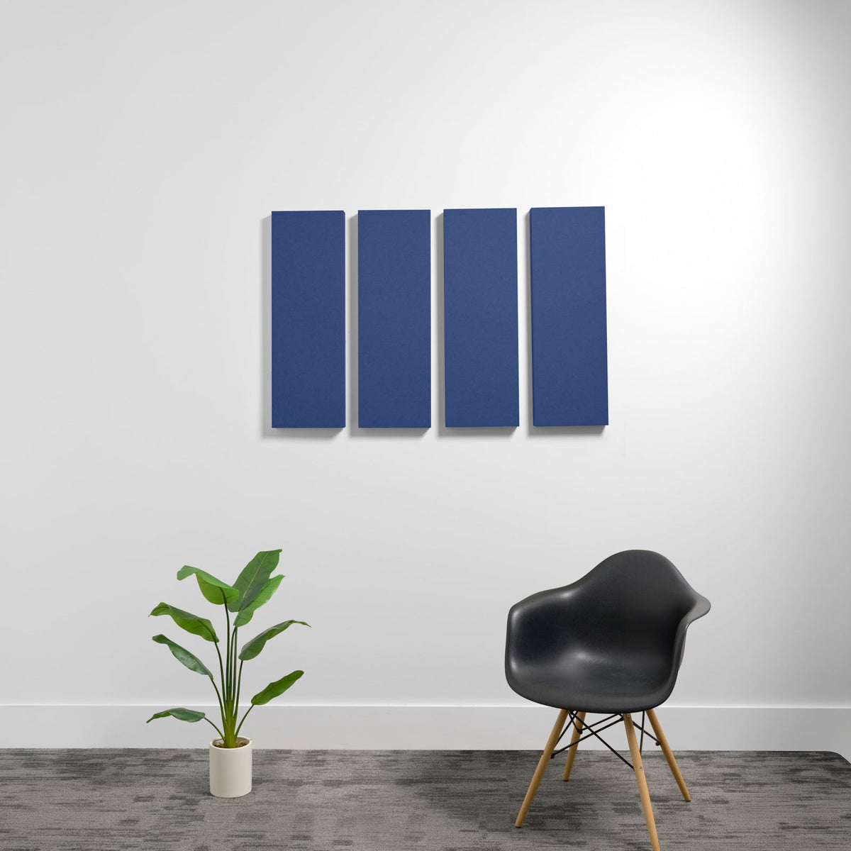 Four dark blue rectangular 3D acoustic panels mounted on a white wall above a black chair and potted plant.